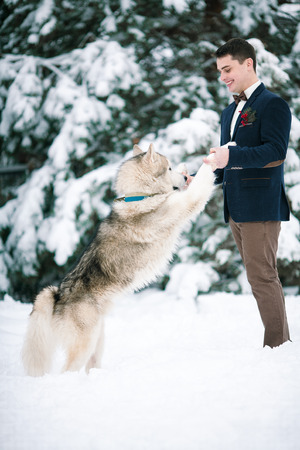 Groom in winter with dog malamute playing in snow. He keeps dog by front paws and smiling.の写真素材