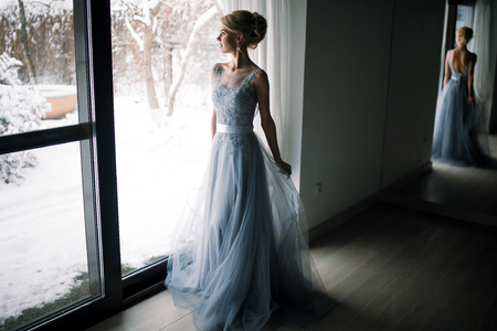 Bride standing in front of big window and looking to snowy garden. Her figure is reflected in large mirror.の写真素材