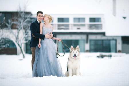 Bride and groom in winter with dog malamute on snow and home background. Groom hugging bride, and she looks at him tenderly.の写真素材