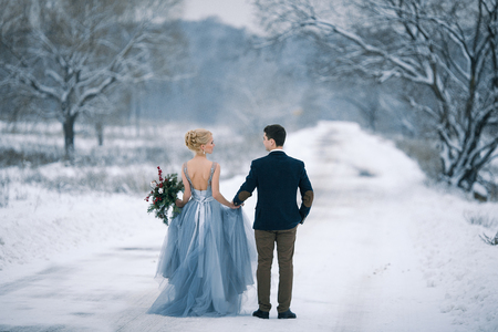 Bride and groom among snowy landscape. Bride and groom are holding hands and walk the snowy road.  View from back. Winter wedding outdoors.の写真素材