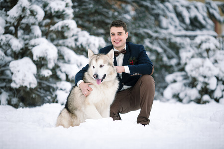 Groom in winter with dog malamute sitting in snow. He hugging dog and smiling.の写真素材