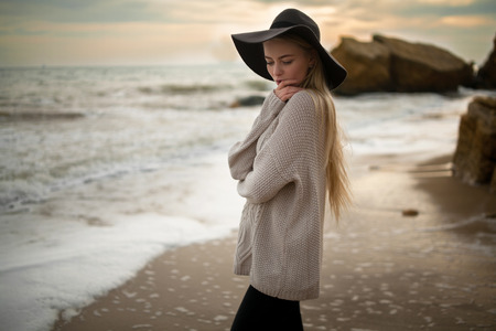 Woman in hat and sweater on winter beach at sunset. She looks pensively at beach. Clouds at sunset sky background and rocks are visible from behind.の写真素材