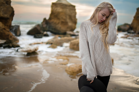 Woman in sweater and hat on winter beach at sunset. She stands and looks down pensively.の写真素材