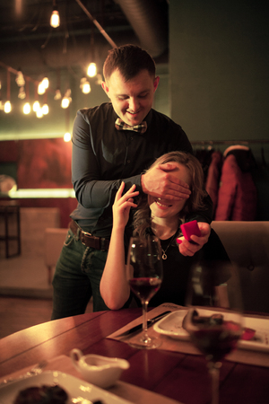 Young man makes woman marriage proposal, he closed her eyes with his hand and holds in front of her jewelry case with engagement ring. Before them is table with food and glasses of wine.の写真素材