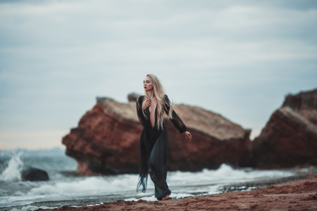Young woman in long transparent black dress among beach and sea. She walks along shore.の写真素材