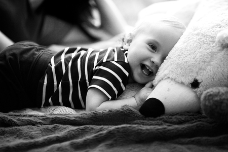 Small baby lies on bed next to teddy bear and laughs. Black and white image.の写真素材