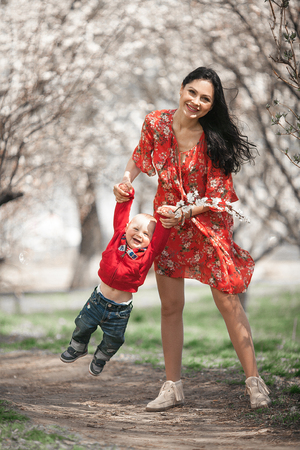 Young mother with her baby on walk in blooming garden. They play and laugh cheerfully among flowering apricot trees.の写真素材