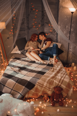 Pregnant woman with her husband sits on bed among lights. They tenderly embrace each other.の写真素材