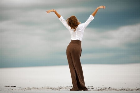 Young woman stands on sand in desert. She is dressed in long skirt and blouse. Back view.の写真素材