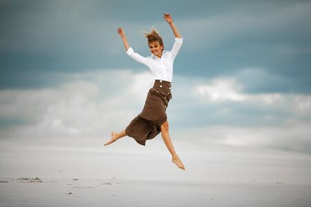 Young woman jumps on sand in desert. She is dressed in long skirt and blouse and joyful laughs.の写真素材