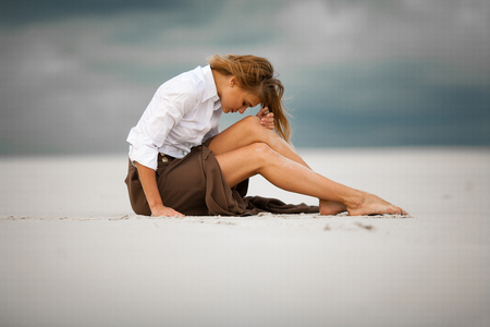 Young sad and pensive woman sits on sand in desert. She is dressed in long skirt and blouse.の写真素材