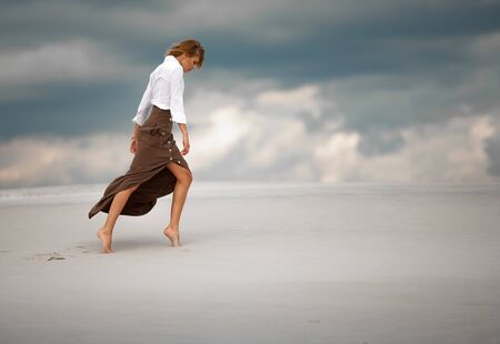Young woman goes barefoot in desert on sky background. She is dressed in long skirt and blouse. Side view.の写真素材