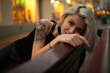 Young woman sits on wet bench on city street in evening. In background there are lights of night city. Portrait.の写真素材