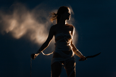 Girl in image of Egyptian mummy stands with metal chain and knife in her hands. She is wrapped in bandages. Around her flies glowing sand in air. Her silhouette illuminatedの写真素材
