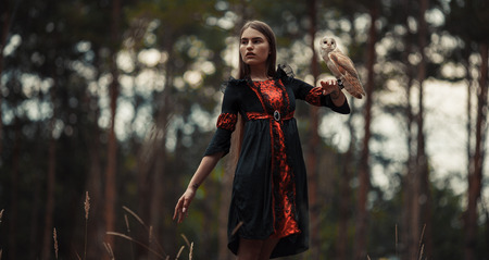 Girl in red and black dress stands with owl on her hand in forest and looks away.の写真素材