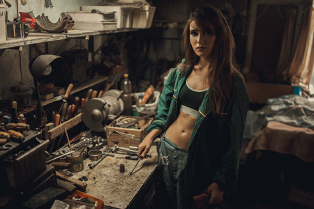 Girl worker in green overalls and t-shirt stands in workshop among tools shelves. She is holding screwdriver.の写真素材