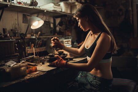 Girl worker in black t-shirt and gloves sits in workshop among tools and unscrews spare part with screwdriver. Nearby is table with lamp and shelves with tools.の写真素材