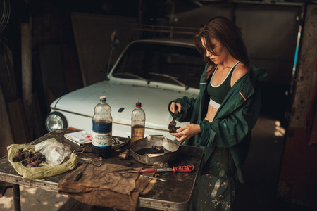 Girl worker in green overalls and t-shirt stands in workshop among tools and lubricates  spare parts from car. In her hands she holds brush, next to her there is bottle of grease.の写真素材