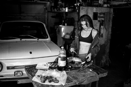 Girl worker in green overalls and t-shirt stands in workshop among tools and lubricates  spare parts from car. In her hands she holds brush, next to her there is bottle of grease. Black and white image.の写真素材