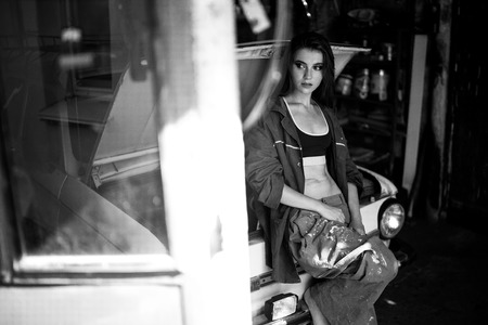 Girl worker in dirty overalls and t-shirt stands in workshop near opened capote of retro car. Black and white image.の写真素材