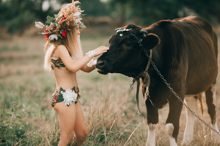 Beautiful little girl in image of nymph with floral head wreath stands on meadow near black cow on chain.の写真素材