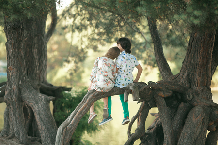 Little girl and boy sit side by side on tree branch and put head on shoulder. First love. Back view.の写真素材