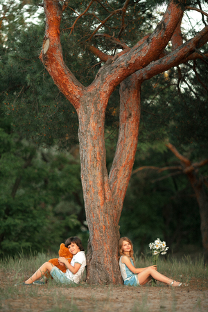 Little girl and boy sit in forest under pine tree. Boy is holding teddy bear in his hands, and girl is flower. First love.の写真素材
