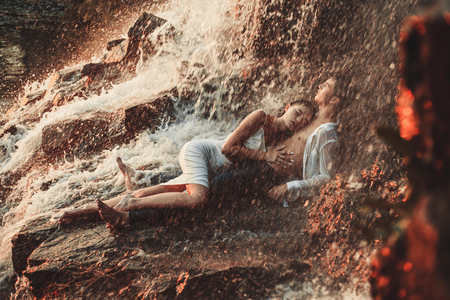 Young enamored wet couple lies on rock and  hugs under spray and drops of waterfall. Around them are visible jets and streams of running water.の写真素材