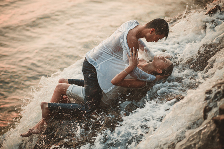 Young enamored wet couple lies on rock and  hugs under spray and drops of waterfall. Around them are visible jets and streams of running water.の写真素材