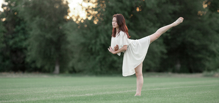 Ballerina in white dress stands and performs swallow pose on lawn and trees background.の写真素材