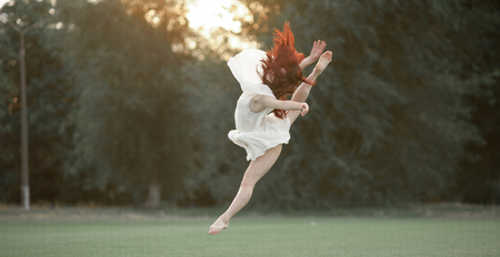 Ballerina in white dress jumping on lawn and trees background.