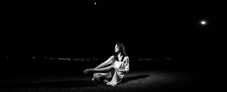 Ballerina in white dress sits on lawn in evening on background of night lights. Black and white image.の写真素材