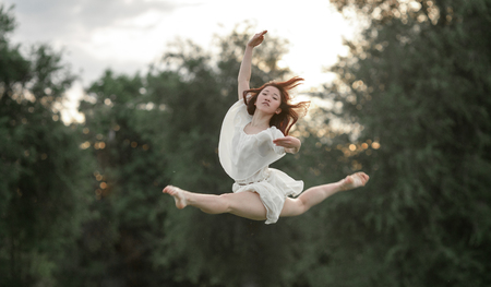 Ballerina in white dress jumps and performs gymnastic twine on trees background.の写真素材