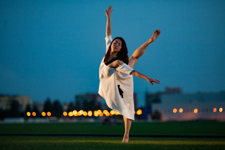 Ballerina in white dress stands and performs swallow pose on lawn and buildings background in evening. Night lights are visible in distance.の写真素材