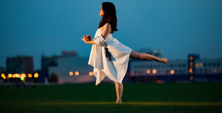 Ballerina in white dress stands and performs swallow pose on lawn and buildings background in evening. Night lights are visible in distance.の写真素材
