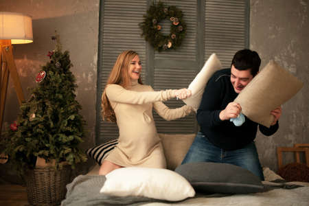 Pregnant woman with her husband before Christmas tree. They are fighting pillows for fun. Light from lamp illuminates room.の写真素材