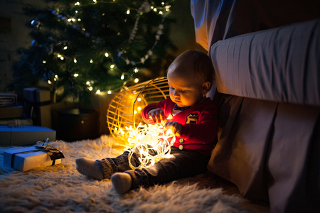 Little baby boy sits on fur rug near Christmas tree, gift boxes and plays by lit garland with lightbulbs.の写真素材