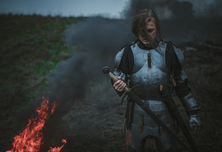 Girl in  armor and with sword in her hands stands on meadow against background of fire, black smoke...