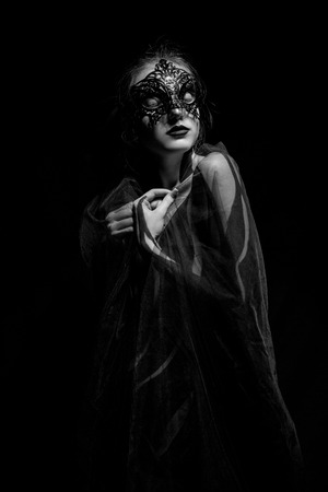 Beautiful young girl wrapped in transparent shawl with painted body and black mask on her face stands against black background. Body art. Black and white image.の写真素材