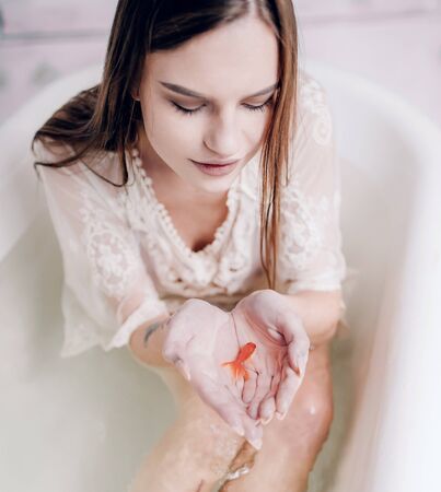 Young woman in lacy dress sits in white bathtub with transparent water and holds goldfish in her palms. Closeup.の写真素材