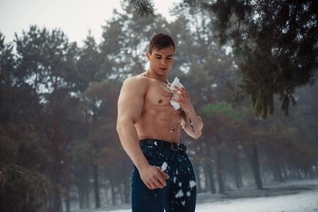 Young bodybuilder with bare torso wipes his body by snow in pine forest in winter. Body hardening...