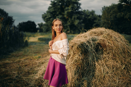 Young smiling woman in shirt and short skirt stands near haystack on meadow.の写真素材