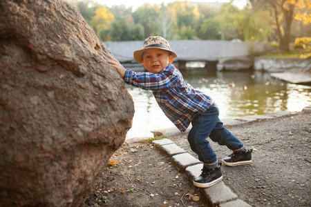 A little boy is playing and pushing a rock during a walk in the park against the background of the lake.の写真素材