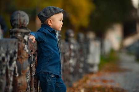 A little boy in cap is standing next to the fence during a walk in the park.の写真素材
