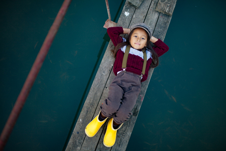 Child girl in cap and yellow rubber boots lies on wooden bridge and smiles on background of river. Top view.の写真素材