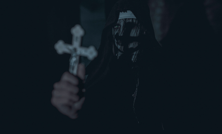 Woman is standing in a darkness with a cross in her hand in a black hooded cloak in an image of a nun possessed by demons. Cosplay.の写真素材