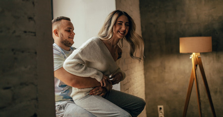 A young couple in love is having fun and laughing joyfully next to the window on background of...