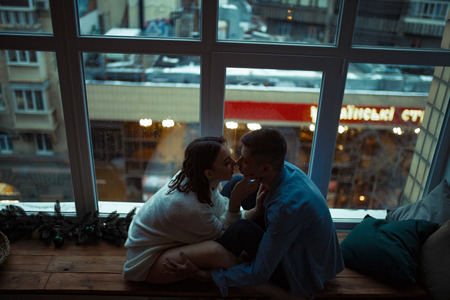 Couple in love sit in a hug on sill next to window against the background of lights of evening city.の写真素材
