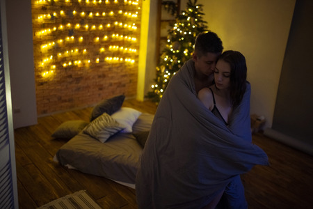 Young lovers are in a hug during foreplay on background of the bed, Christmas tree and decorations in dark room.の写真素材
