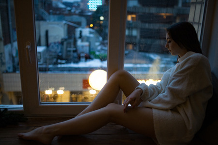 A young woman sits on the windowsill and looks through the glass with raindrops at the lights of the evening city.の写真素材